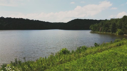 A serene lake surrounded by lush greenery and rolling hills under a partly cloudy sky.