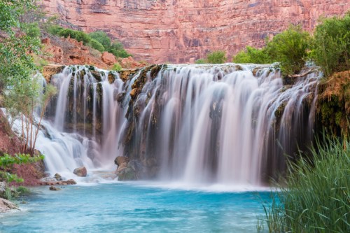 A serene waterfall cascades into a turquoise pool, surrounded by lush greenery and red rock formations.