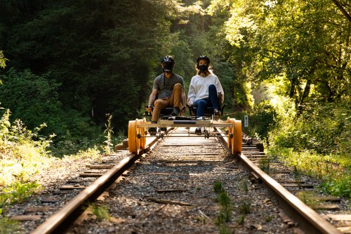 Two people ride a rail bike on a forested train track, surrounded by greenery and sunlight.