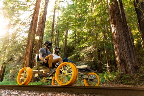 Two people wearing helmets sit on a unique, wheeled vehicle on a forest path surrounded by tall trees.