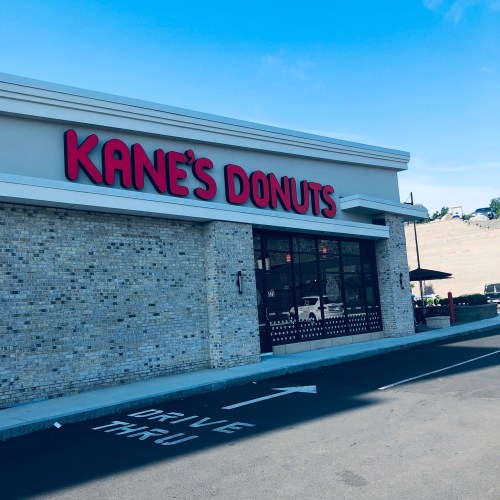 Exterior of Kane's Donuts shop with a clear blue sky and a drive-thru lane visible in front.