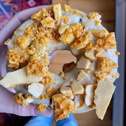 A hand holding a donut topped with caramelized honeycomb and crunchy pieces on a light glaze.