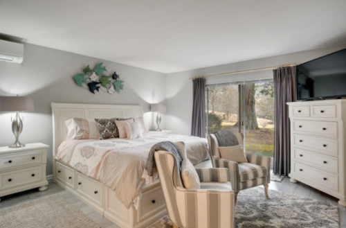Cozy bedroom with a white bed, striped chair, dresser, and large window overlooking a serene outdoor view.