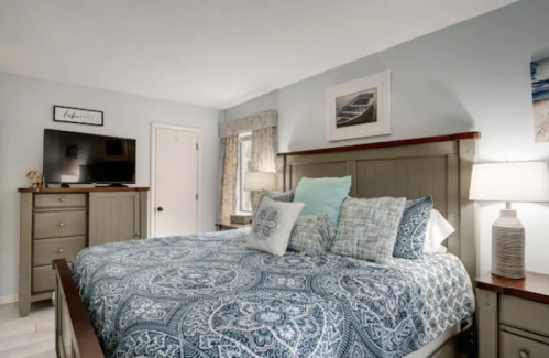 Cozy bedroom with a patterned blue bedspread, nightstands, lamps, and a TV on a dresser against light blue walls.