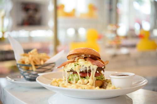 A gourmet burger topped with bacon and cheese, served with a side of fries and dipping sauce on a white plate.