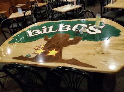 A wooden table with a painted tree design and the name "Bilbo's" in bold letters, surrounded by black chairs.