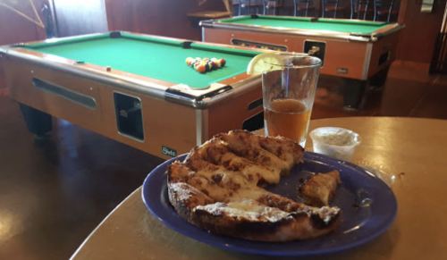 A plate of grilled chicken with a drink, set in a bar with a pool table in the background.