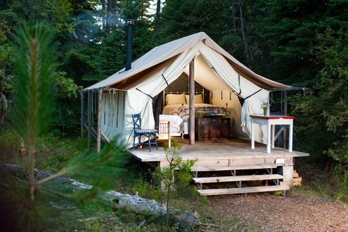 A cozy glamping tent on a wooden platform, surrounded by trees, featuring a bed and a small table outside.