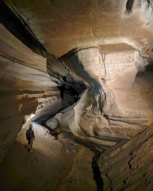 A person stands in a narrow, illuminated cave with smooth, layered rock formations and a small stream.