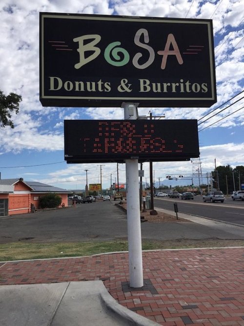 Sign for Bosa Donuts & Burritos with a digital display, set against a cloudy sky and roadside view.