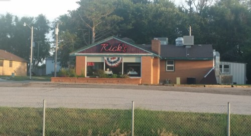 A brick building with a sign reading "Rick's" and an "Open" banner, surrounded by trees and a grassy area.