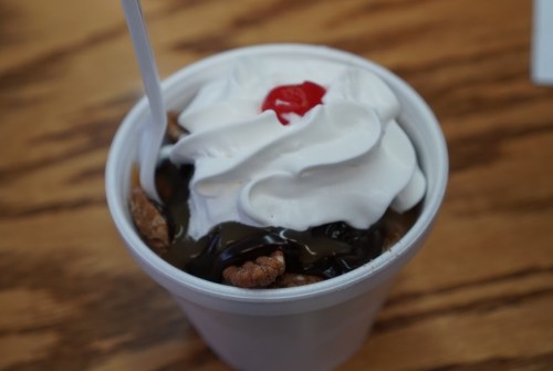 A cup of dessert topped with whipped cream, chocolate sauce, and a cherry, resting on a wooden surface.