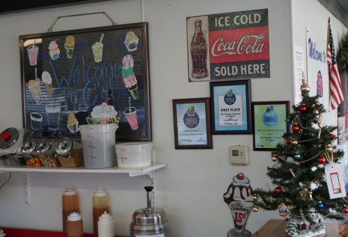 A cozy ice cream shop interior with a welcome sign, Coca-Cola advertisement, awards, and a small Christmas tree.