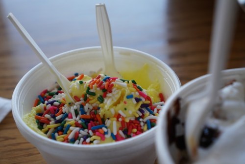 A cup of yellow ice cream topped with colorful sprinkles and two plastic spoons, set on a wooden table.