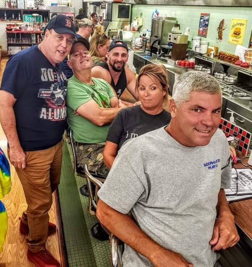 A group of five people sitting at a diner counter, smiling and chatting in a casual, lively atmosphere.