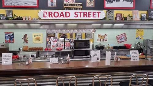 A retro diner counter with a "Broad Street" sign, vintage decor, and a menu featuring hot dogs and burgers.