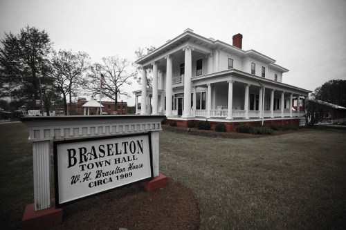 Historic Braselton Town Hall, a white-columned house from 1909, surrounded by green grass and trees on a cloudy day.