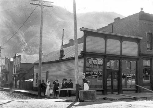 Historic black-and-white photo of a street scene with people in front of a Brunswick store and wooden buildings.