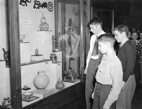 Three boys observe artifacts in a museum display case, with various objects and sculptures visible behind glass.