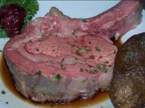 A juicy slice of prime rib with herbs, served alongside a baked potato and a small portion of sauce.