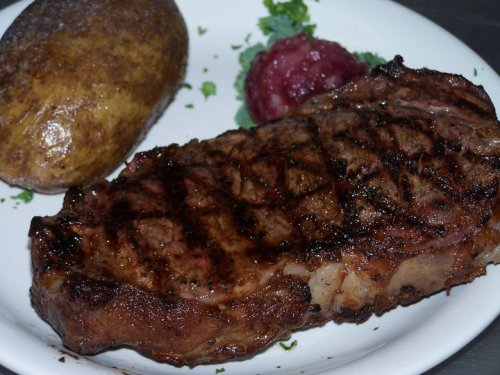 A grilled steak on a plate, accompanied by a baked potato and a small serving of red sauce.