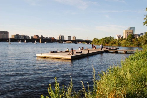 A wooden dock extends into a calm river, with people relaxing and sailboats in the distance against a city skyline.