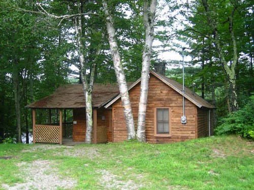 A rustic wooden cabin surrounded by trees, with a porch and grassy area in front.