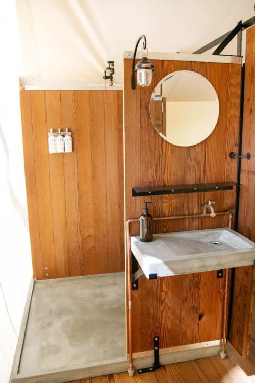 A modern bathroom with wooden walls, a circular mirror, a concrete sink, and a shower area.