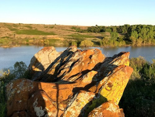 Sunlit rocky outcrop in the foreground with a calm river and lush greenery in the background under a clear blue sky.
