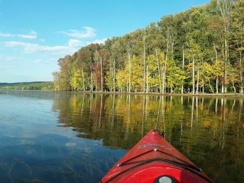 A red kayak on calm water, reflecting colorful trees along the shore under a clear blue sky.