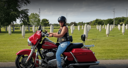 A woman in a helmet rides a red motorcycle on a road lined with large, white corn-shaped sculptures in the background.