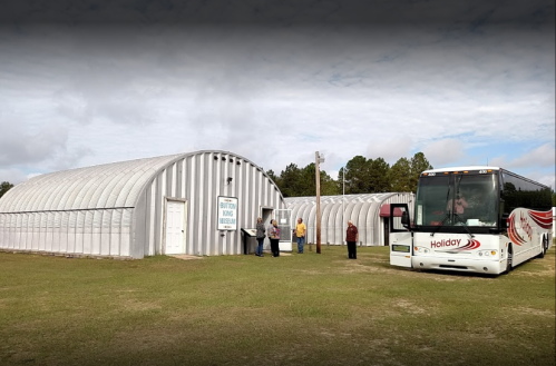 A bus parked outside a large metal building with people gathered near the entrance on a grassy area.