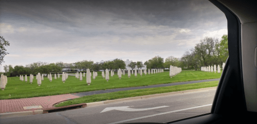 A view of a cemetery with numerous white headstones on a grassy field, seen from a vehicle window.