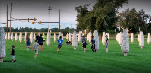 Children and adults play among large corn-shaped sculptures in a grassy field at dusk.