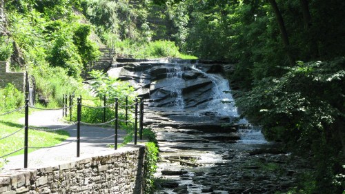 A serene waterfall cascades over rocky steps, surrounded by lush greenery and a winding path.
