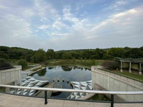 A serene landscape featuring a reflective pond surrounded by greenery and modern architectural elements.