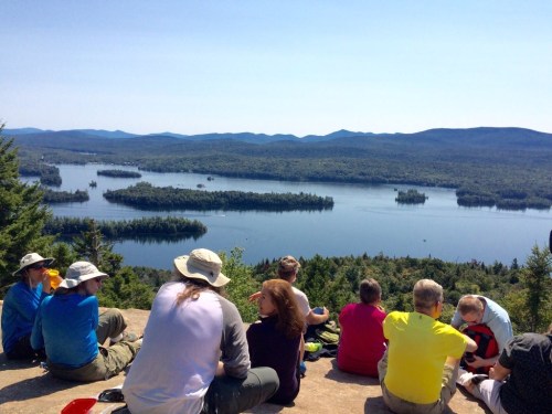 A group of hikers sits on a rocky ledge, overlooking a serene lake surrounded by forested mountains on a sunny day.
