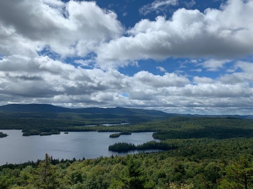 A scenic view of a lake surrounded by lush green forests and mountains under a partly cloudy sky.