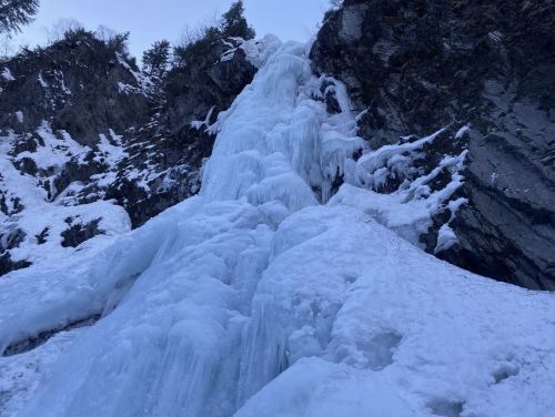 A frozen waterfall cascading down rocky terrain, surrounded by snow and ice.