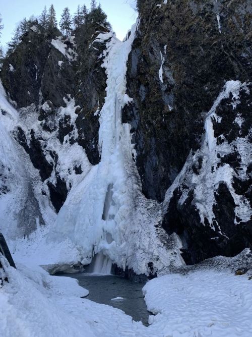 A frozen waterfall cascading down rocky cliffs, surrounded by snow-covered terrain and a small river below.