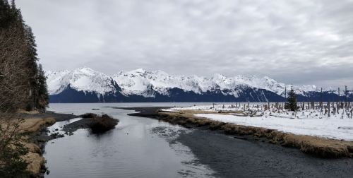 A serene landscape featuring a river winding through snow-covered terrain, with majestic mountains in the background under a cloudy sky.