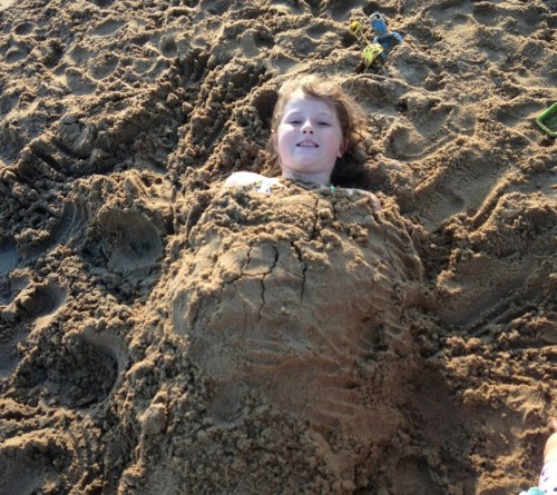A child lying on the beach, covered in sand, with a playful expression and toys scattered nearby.