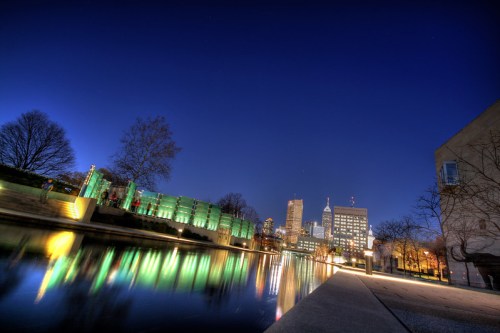 City skyline at dusk, reflecting in a calm river, with illuminated buildings and trees along the waterfront.