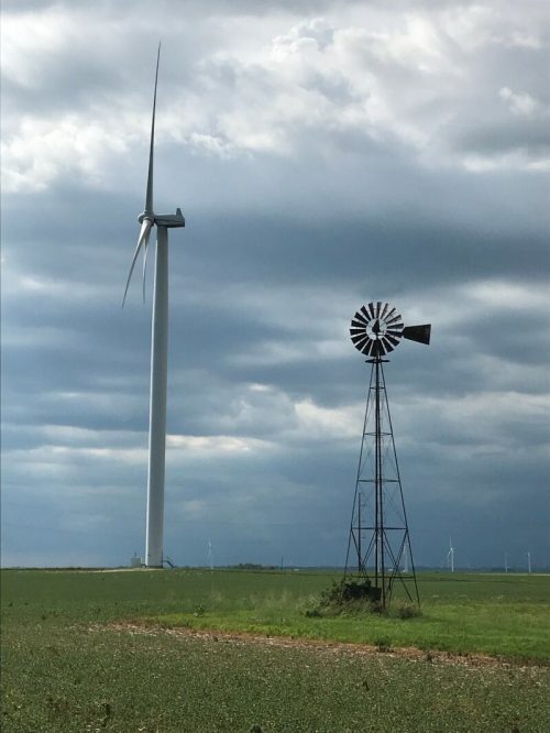 A modern wind turbine stands next to a traditional windmill in a green field under a cloudy sky.