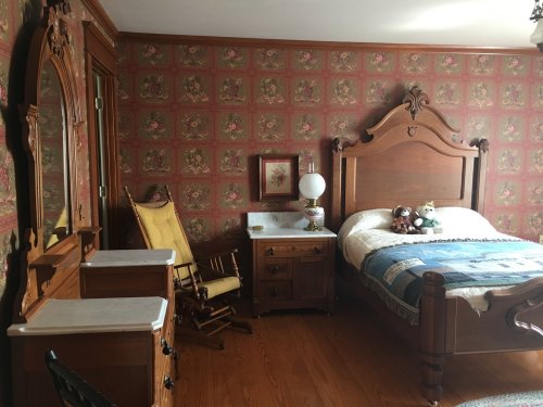 A vintage bedroom with floral wallpaper, a wooden bed, a rocking chair, and a dresser, featuring stuffed animals on the bed.