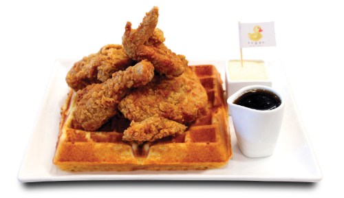 A plate of crispy fried chicken on a waffle, served with syrup and a small cup of dipping sauce.