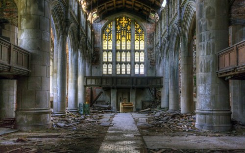 An abandoned church interior with tall stone columns, broken windows, and debris scattered on the floor.