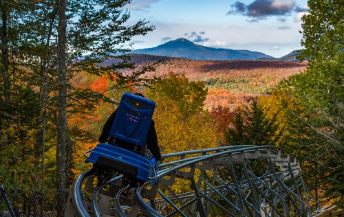 A person rides a blue coaster on a track with vibrant autumn foliage and mountains in the background.