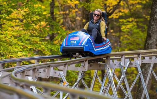 A woman rides a blue alpine coaster through a colorful autumn forest.