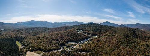 Aerial view of a winding road through colorful autumn forests and mountains under a clear blue sky.